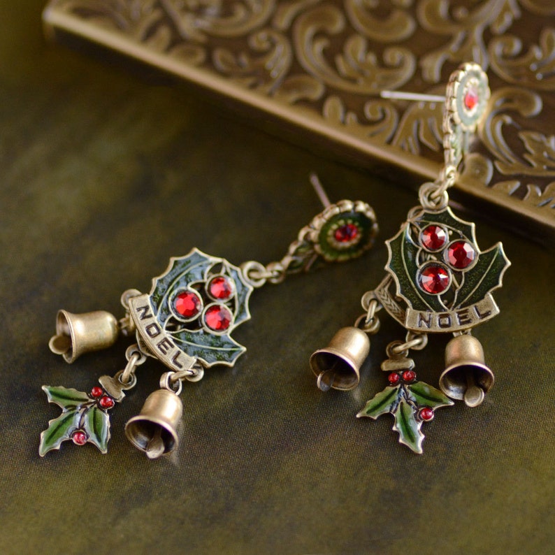 May include: A pair of gold-tone dangle earrings with a green holly leaf design, red rhinestone accents, and the word "NOEL" on each earring. The earrings also feature three small bells.