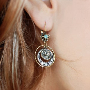 May include: Gold tone dangle earrings with a circular design featuring a small, light blue gemstone and a coin-shaped charm with a zodiac symbol.