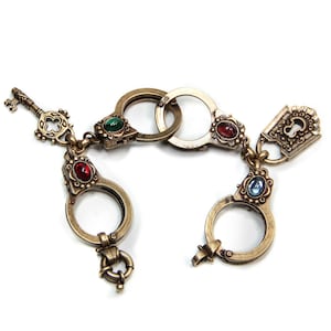 Puede incluir: Un brazalete dorado con múltiples dijes en forma de ojo de cerradura, cada uno adornado con una gema de color diferente. Los dijes están conectados por una cadena e incluyen un pequeño dije de llave.