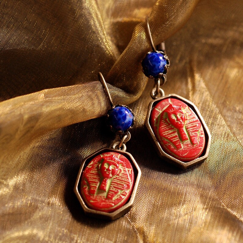May include: A pair of red and blue earrings with a carved pharaoh design. The earrings are made of metal and have a blue glass bead at the top.