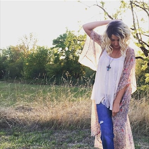 May include: A woman wearing a floral print kimono over a white tank top and blue jeans. She is standing in a field with tall grass and trees in the background. She is wearing a silver necklace with a turquoise cross pendant.