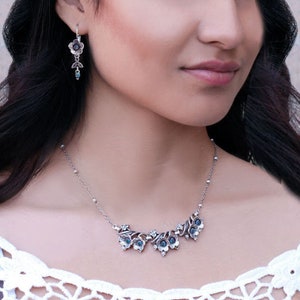 Op de afbeelding: Een zilveren ketting met een delicate ketting en een bloemvormige hanger met blauwe edelstenen. De hanger bestaat uit verschillende kleine bloemen die met wijnstokken zijn verbonden.