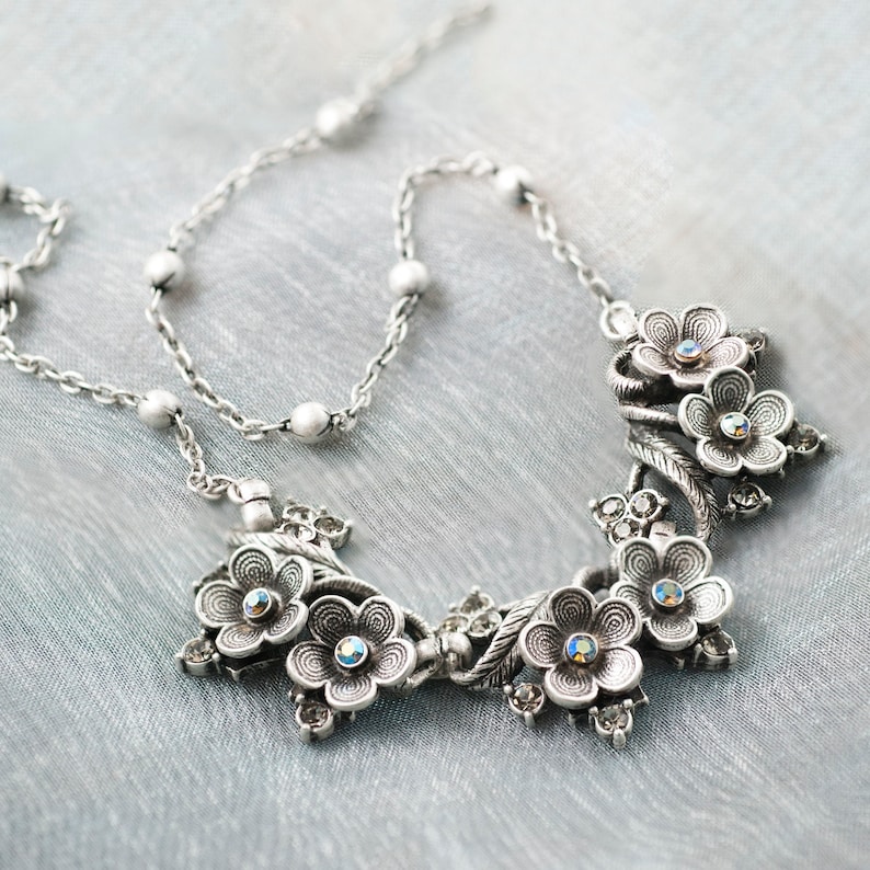 Op de afbeelding: Een zilveren ketting met een bloemmotief met meerdere bloemen met heldere strass steentjes. De ketting is een eenvoudige zilveren ketting met kleine ronde kralen.