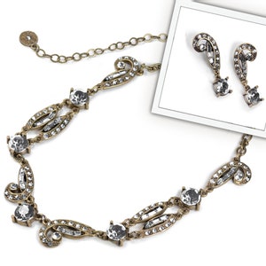 May include: A gold tone necklace and earrings set with clear crystal accents. The necklace features a delicate chain and a repeating pattern of crystal-encrusted swirls and leaves. The earrings are drop style with a similar design.