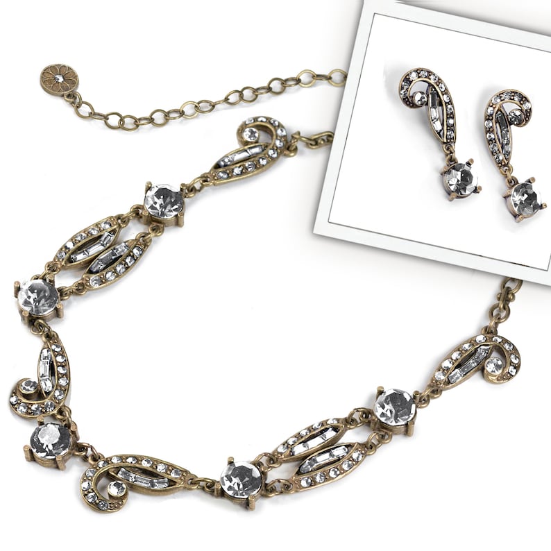 May include: A gold tone necklace and earrings set with clear crystal accents. The necklace features a delicate chain and a repeating pattern of crystal-encrusted swirls and leaves. The earrings are drop style with a similar design.
