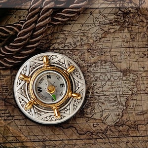 May include: A silver compass with a gold-coloured face and a compass needle pointing north. The compass is surrounded by a decorative design and is sitting on top of an old map.