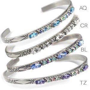 May include: Four silver bracelets with different colored crystals. The bracelets are stacked on top of each other. The crystals are in the following colors: aqua, clear, blue, and topaz. The bracelets are labeled with the corresponding crystal color: AQ, CR, BL, TZ.