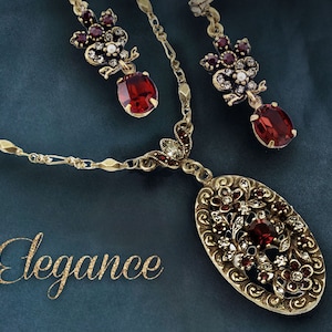 May include: A gold necklace with a red and white floral pendant and matching earrings. The necklace features a delicate chain and a large oval pendant with intricate details. The earrings are small and feature a cluster of red and white stones.