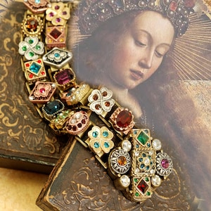 May include: A gold-toned bracelet with a multi-colored design featuring various gemstones and clover charms. The bracelet is on a brown surface with a painting of a woman in the background.