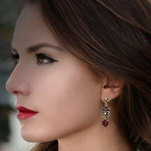 May include: A pair of dangle earrings with a red gemstone and a cluster of smaller red and clear gemstones. The earrings are gold-toned.