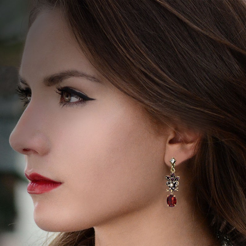 May include: A pair of dangle earrings with a red gemstone and a cluster of smaller red and clear gemstones. The earrings are gold-toned.