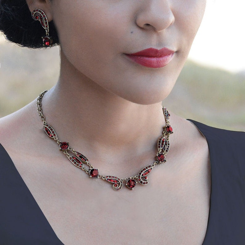 May include: A gold necklace with a delicate design featuring red gemstone accents. The necklace has a leaf-like pattern and is adorned with round red stones.