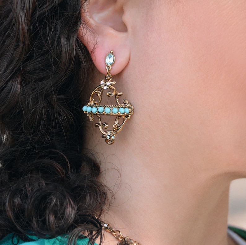 May include: A pair of gold filigree earrings with a row of turquoise blue beads. The earrings are dangling from the earlobe.