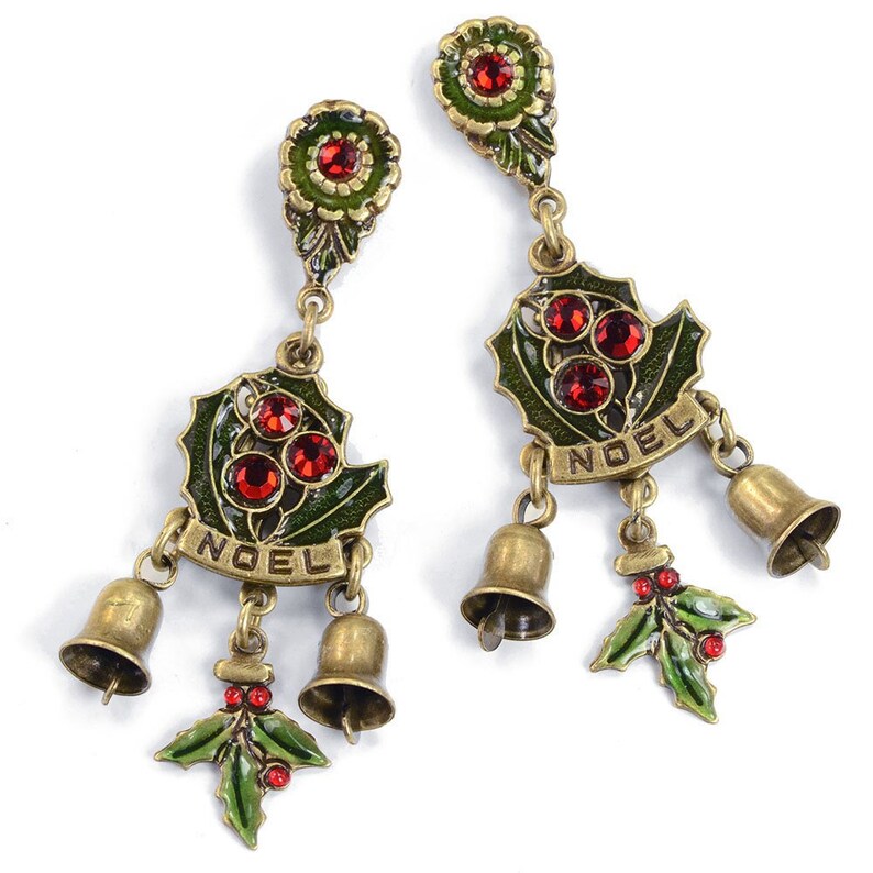 May include: A pair of dangle earrings with a green holly leaf design, red crystals, and gold bells. The earrings have the word "NOEL" written on them.