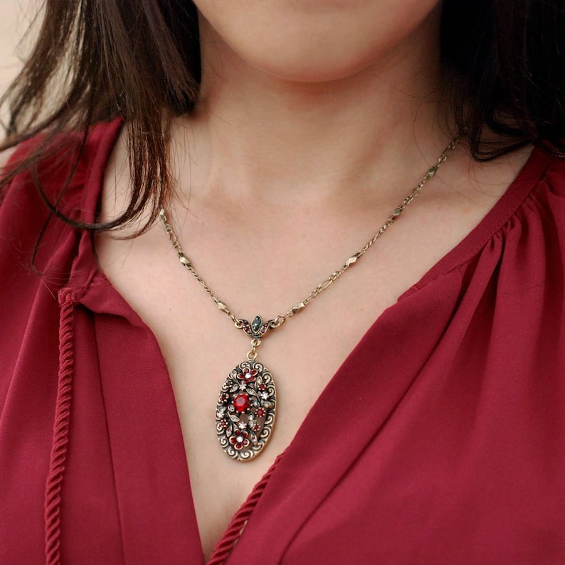 May include: A gold chain necklace with a red and clear crystal pendant. The pendant is oval shaped and features a floral design.