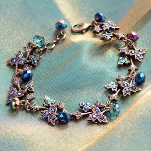 May include: A delicate bracelet featuring a leafy design with blue and iridescent crystals. The bracelet is made of metal and has a lobster claw clasp.