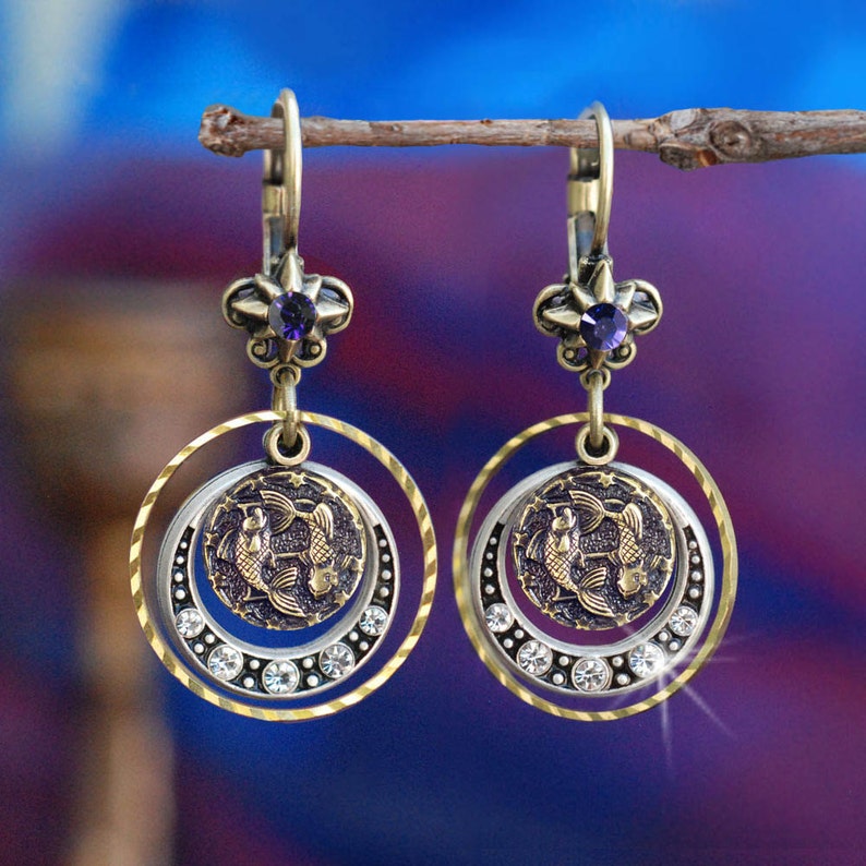 May include: Gold tone earrings with a purple gemstone and a silver circle with a gold fish design. The circle is surrounded by clear rhinestones.