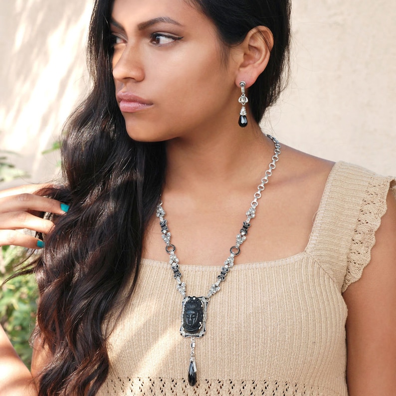 May include: A silver chain necklace with a black stone pendant featuring a carved face. The pendant is attached to a black teardrop-shaped stone. The necklace is worn by a person with long black hair.
