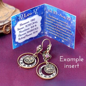 May include: Gold tone earrings with a pink stone and a circular design featuring the astrological symbol for Leo. The earrings are accompanied by a card with the text "Leo" and information about the astrological sign, including its element, stone, numbers, flower, and ruling planet. The text on the card reads "Jul. 23 - Aug. 22 Exuberant, principled, honorable, intelligent, brave, capable, ambitious, generous, extravagant, disciplined, proud, trusting, stylish, committed, deliberate, egocentric, witty, opinionated, energetic."