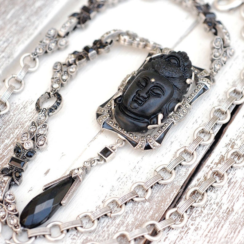 May include: A silver chain necklace with a black carved Buddha pendant. The pendant is set in a silver frame with black enamel accents. The chain is made of small silver links.
