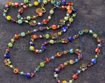 May include: A long, multi-strand necklace with colorful glass beads and gold accents. The beads are a mix of round and square shapes, and the necklace is strung on a gold chain.