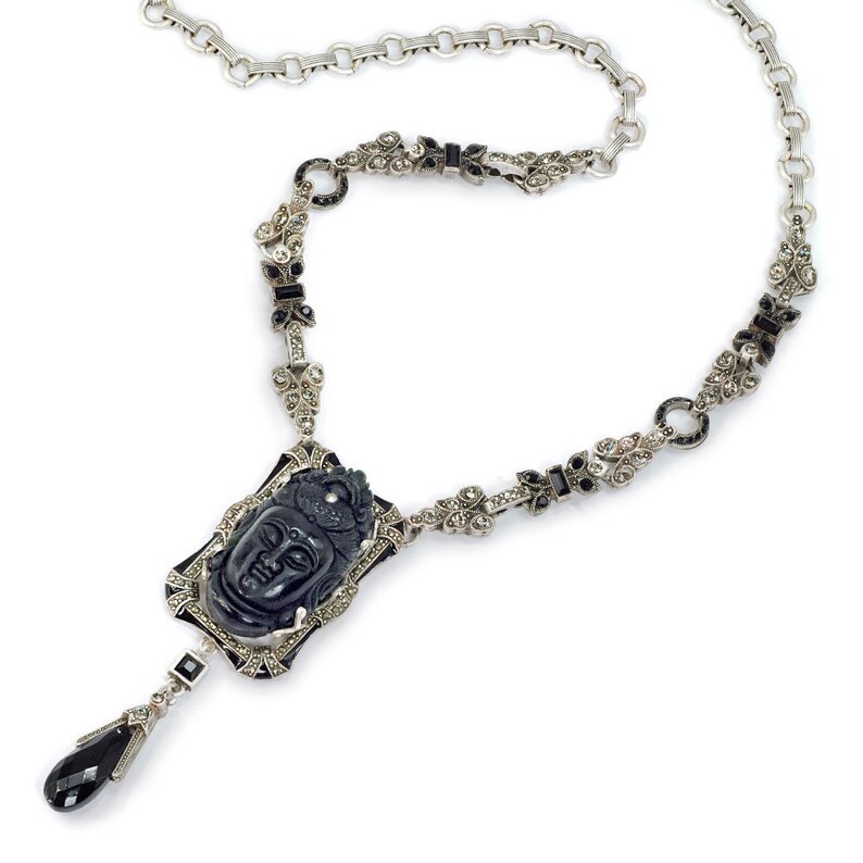May include: A silver necklace with a black onyx pendant featuring a carved Buddha face. The necklace is adorned with black and white marcasite stones.