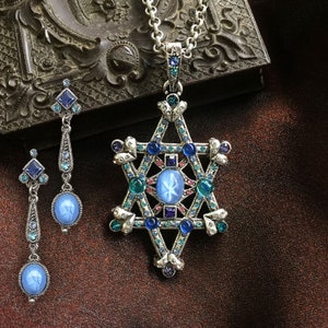 May include: A silver-toned jewelry set featuring a Star of David pendant necklace and matching earrings. The pendant and earrings are adorned with blue and teal gemstones and a central oval blue stone. The set is displayed on a brown surface.