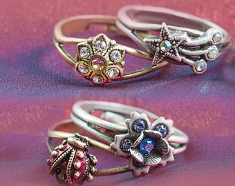 May include: Three silver rings with decorative accents. The top ring features a star with a blue rhinestone and a silver design. The middle ring has a flower with clear rhinestones and a gold band. The bottom ring has a flower with blue rhinestones and a ladybug with red and black accents.