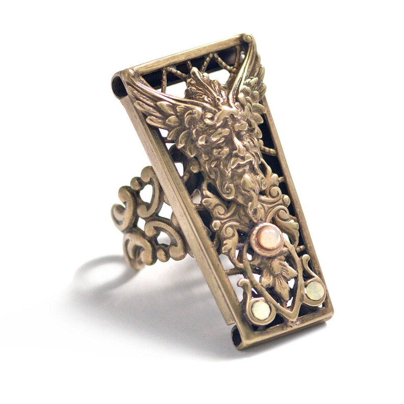 May include: A gold-toned ring with a detailed design featuring a winged creature with a human face and opalescent stones. The ring is made of metal and has a filigree design.
