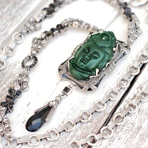 May include: A silver chain necklace with a green jade Buddha pendant and black accents. The pendant is set in a silver frame with black enamel details. The chain is made of silver and has a black onyx drop.