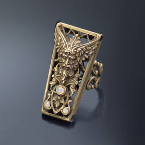 May include: A gold-toned ring with a detailed design featuring a green-faced figure with wings and opalescent stones. The ring is made of metal and has a rectangular shape.