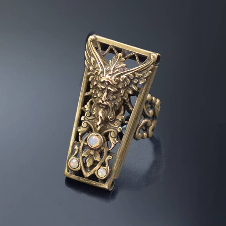 May include: A gold-toned ring with a detailed design featuring a green-faced figure with wings and opalescent stones. The ring is made of metal and has a rectangular shape.