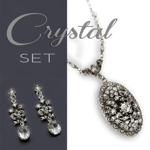May include: A silver crystal necklace and earrings set. The necklace features a large oval pendant with a floral design and clear crystals. The earrings are drop earrings with a similar floral design and clear crystals.