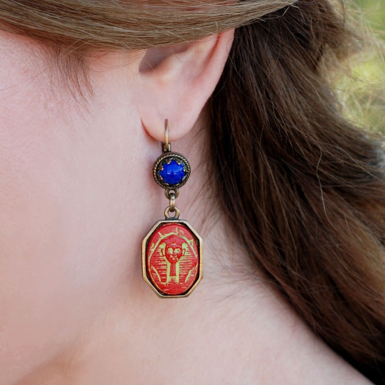 May include: Red and gold Egyptian-style earrings with a blue gemstone accent. The earrings are made of metal and have a dangling design.
