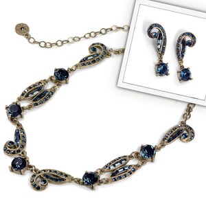 May include: A gold necklace and earrings set with a blue crystal design. The necklace features a delicate chain with a pendant that has a swirling pattern and blue crystals. The earrings are drop earrings with a similar swirling pattern and blue crystals.