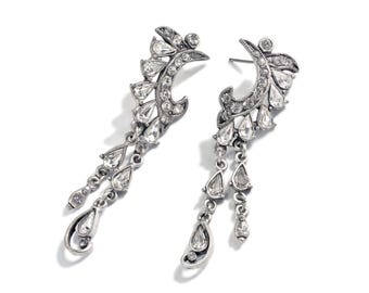May include: A pair of silver dangle earrings with crystal accents. The earrings feature a crescent moon design with a teardrop-shaped crystal dangle.