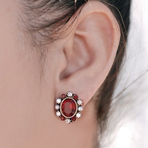 Puede incluir: Un par de pendientes de piedras preciosas rojas y blancas con un dise&ntilde;o floral. Los pendientes presentan una piedra roja grande rodeada de piedras blancas m&aacute;s peque&ntilde;as.