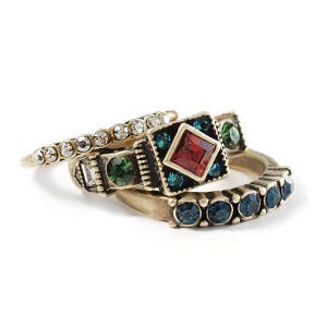 May include: A collection of gold-toned rings with various gemstone embellishments. One ring features a row of clear crystals, while others showcase emerald green, ruby red, and sapphire blue stones. The rings are stacked together, creating a layered look.