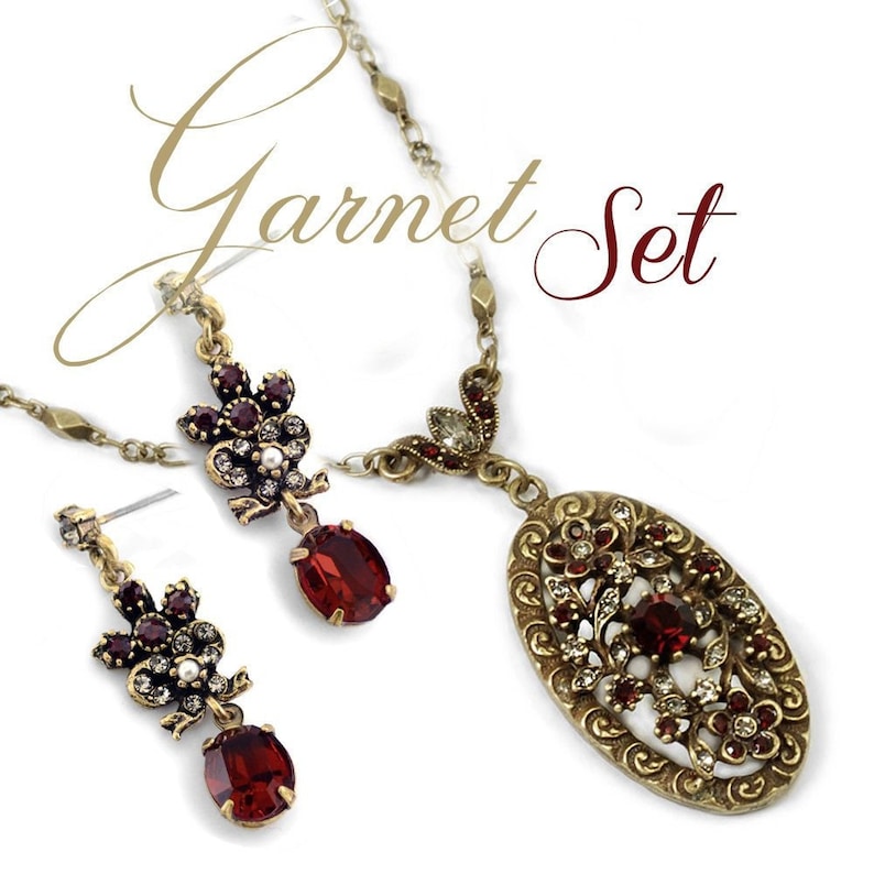 May include: A gold necklace and earrings set with a red garnet stone pendant and matching earrings. The pendant features a floral design with a red garnet stone in the center. The earrings are also floral with a red garnet stone at the bottom.