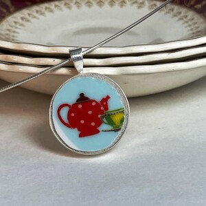 Può includere: Collana con pendente in argento con un ciondolo rotondo. Il ciondolo presenta una teiera rossa a pois bianchi e una tazza da tè verde su sfondo azzurro chiaro. La collana ha una catena sottile.