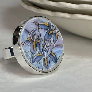May include: A silver ring with a round, floral design. The ring features a detailed image of blue irises with yellow accents on a light purple background. The ring's band is a simple, polished silver.
