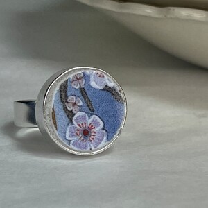 May include: A silver ring with a round, floral design. The ring features a light blue background with delicate purple and white flowers. The ring's band is a simple, polished silver.