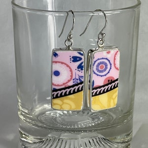 Può includere: Un paio di orecchini rettangolari con un design floreale rosa, giallo e blu. Gli orecchini hanno ganci color argento e sono esposti in un bicchiere trasparente. Gli orecchini hanno un motivo unico e colorato.