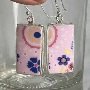 Può includere: Un paio di orecchini rettangolari con un motivo floreale rosa. Gli orecchini hanno montature e ganci per le orecchie in metallo argentato. Il motivo floreale comprende fiori blu, gialli e bianchi su sfondo rosa.