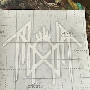 May include: A white vinyl decal featuring a stylised, symmetrical emblem with sharp, angular lines and a central crown-like motif, applied to a clear Cricut transfer tape sheet with a printed grid pattern and the text Cricut visible.
