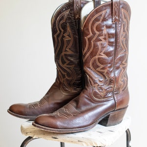 May include: A pair of brown leather cowboy boots with intricate stitching detail. The boots are sitting on a white stool with black metal legs.