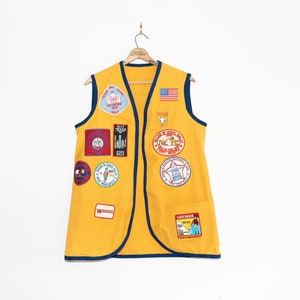 May include: A yellow sleeveless vest with blue trim and a variety of patches sewn on. The patches feature images of a cowboy, a bull, a star, and a map of Texas. The vest has a pocket on the front with a white label that says "Wrangler".