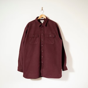 Może przedstawiać: Bordowa koszula zapinana na guziki z dwiema kieszeniami na piersi, wisząca na drewnianym wieszaku. Koszula ma kołnierzyk i długie rękawy. Tkanina wydaje się być miękkim, szczotkowanym materiałem.