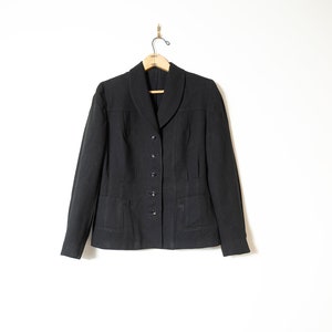 Peut inclure: Un blazer noir avec un col cranté, une fermeture à boutons et deux poches. Le blazer est suspendu à un cintre en bois sur un fond blanc.