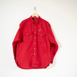 Może przedstawiać: Czerwona koszula z długim rękawem zapinana na guziki, z dwoma dużymi kieszeniami z przodu. Koszula ma małe haftowane logo na lewej piersi.